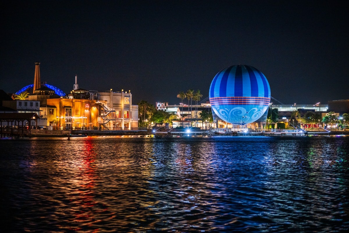 The Disney Springs waterfront at night, seen across Village Lake: the Aerophile balloon glowing over the warmly lit buildings of The Landing and the West Side, their red, white and blue lights reflected on the water.