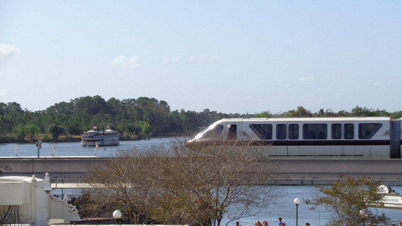 A monorail passing beside Seven Seas Lagoon with a ferry on the water behind it